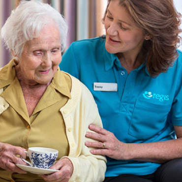 An aged care worker helps an older woman drink tea.